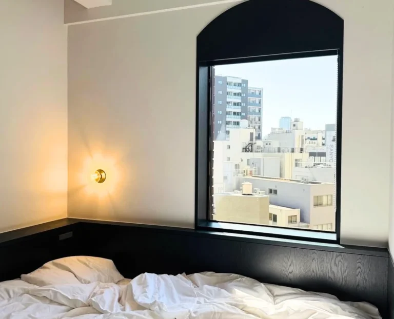 Minimal boutique hotel bedroom with soft natural light and city view through an arched window, calm luxury interior design and quiet travel stay concept.