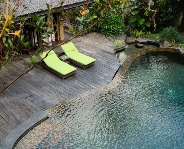Tropical boutique hotel pool surrounded by lush greenery and wooden deck lounge chairs, peaceful luxury resort atmosphere in nature.